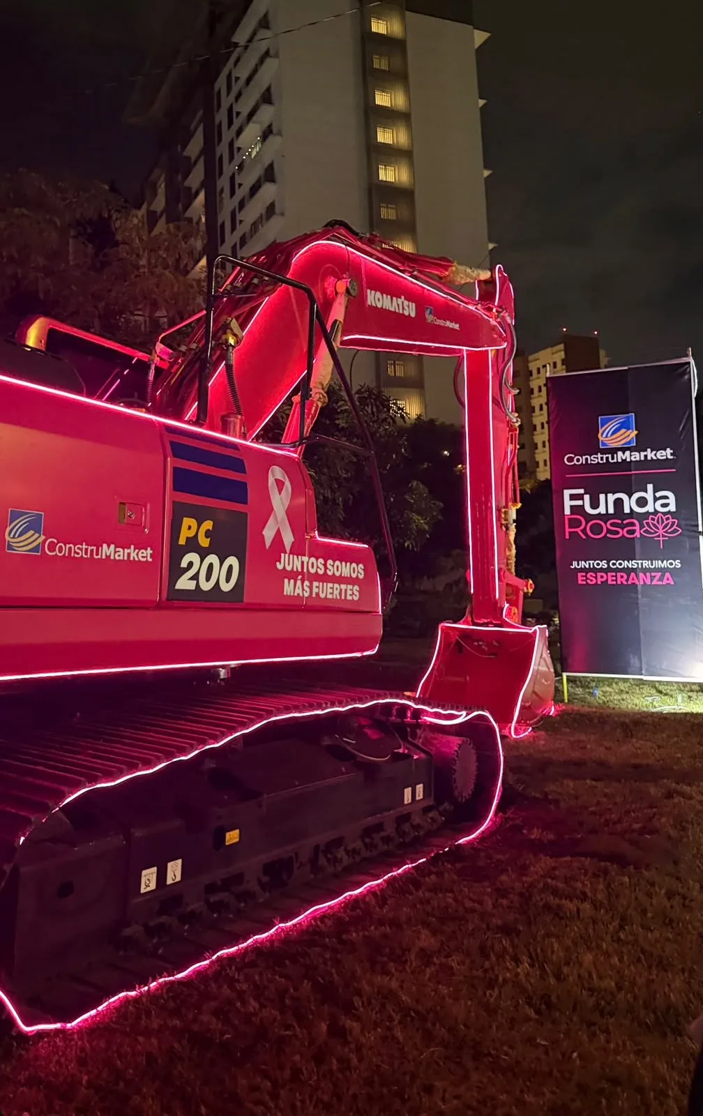 Komatsu PC200 excavator lit at night with pink light strips, next to the FundaRosa banner